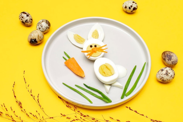 Ostern lustig kreative gesunde Frühstück Mittagessen Essen Idee für Kinder Kinder Hase aus gekochten Hühnereiern geschälte Karotten Grüns auf Platte gelber Tischhintergrund Ansicht von oben flach liegen