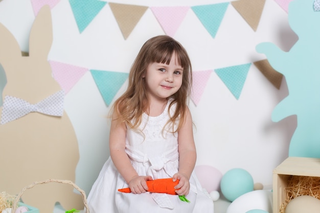 Ostern! Porträt eines schönen kleinen Mädchens in einem weißen Kleid, das eine Karotte hält. Ostern Innendekoration, Frühlingsdekor. Familienurlaub, Traditionen. Kleiner Bauer. Ernte. Vegetarisch, Gemüse