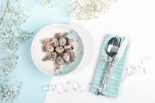 Ostertischgedeckungskonzept mit Platte verzierte Frühlingsblumen mit Wachteleiern, sanften weißen Blumen, Besteck auf Serviette auf weißem und hellblauem Hintergrund.