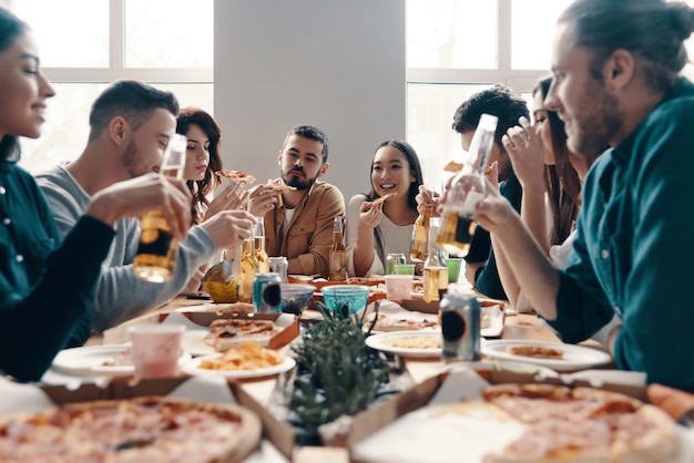 Ótima festa. grupo de jovens em trajes casuais comendo pizza e sorrindo durante um jantar dentro de casa