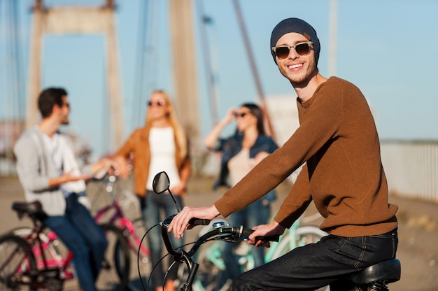 Ótimo dia com melhores amigos. jovem bonito de bicicleta olhando para a câmera e sorrindo enquanto seus amigos conversando ao fundo