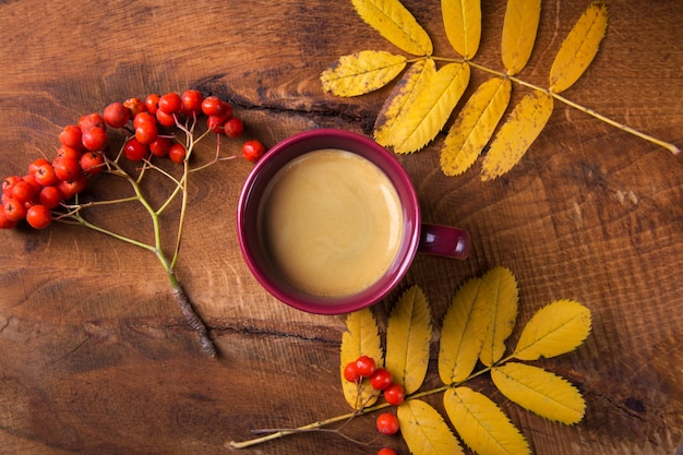 Otoño, hojas y bayas de serbal, una taza de café humeante en una mesa de madera Vista superior.