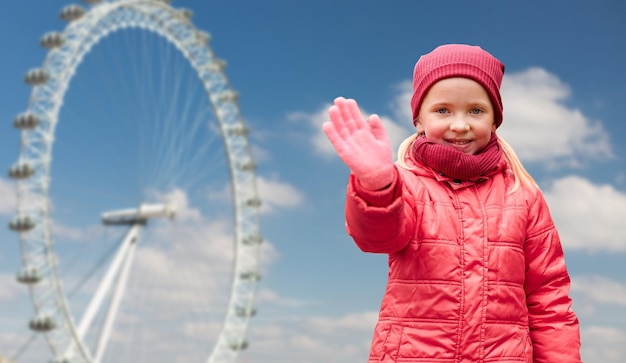 otoño, infancia, gesto, naturaleza y concepto de la gente - niña feliz agitando la mano sobre la rueda del ferry de Londres y el fondo del cielo azul