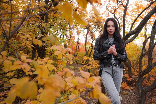 Otoño joven mujer caucásica con una chaqueta de cuero negro y un suéter gris y jeans en el parque de otoño. Clima cálido.