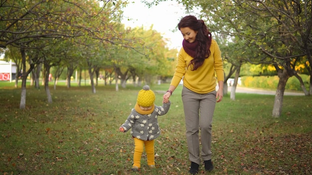 Otoño positivo. Mamá e hija vestidas con hermosas ropas de moda caminan por el parque sobre las hojas amarillas caídas en un cálido día soleado