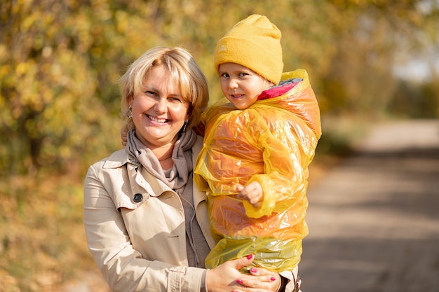 Otoño retrato de madre e hija vestida con sombrero amarillo, mamá sosteniendo a la niña en sus brazos entre hojas amarillas