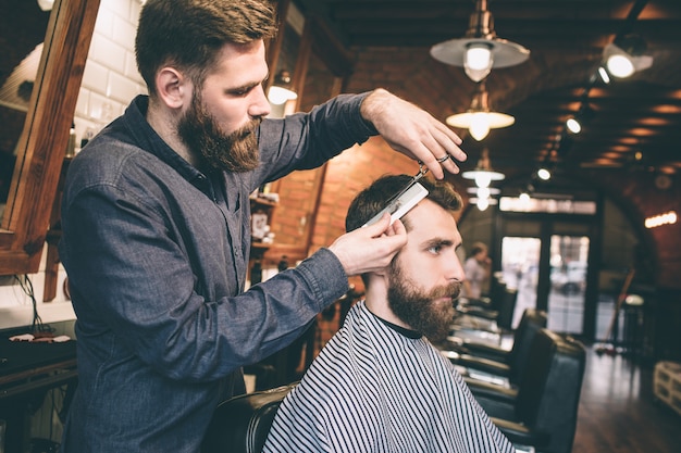 Otra foto de tipos barbudos en la barbería. Un hombre está cortando el pelo de otro hombre. El procedimiento es muy largo pero intenso.