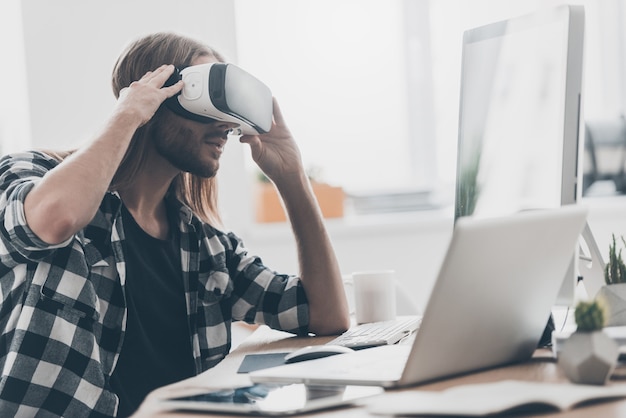 ¡Otra realidad está aquí! Apuesto joven con cabello largo ajustando su casco de realidad virtual mientras está sentado en su escritorio en la oficina creativa