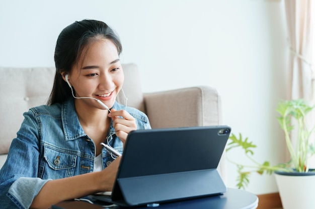 Oung asiatische Frauen führen Videokonferenzen mit ihrer Kollegin mit Tablet-Computer über Technologie zu Hause durch. Schöne Frau, die ein Jeanshemd trägt, spricht über die Kopfhörer, die von zu Hause aus arbeiten