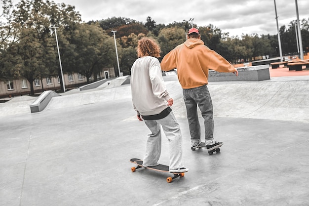 Outdoor-Aktivitäten. Rückansicht von Mann und Mädchen in Jeans und Sweatshirt, die tagsüber auf Skateboards auf einer ebenen Fläche im Skatepark nebeneinander gleiten