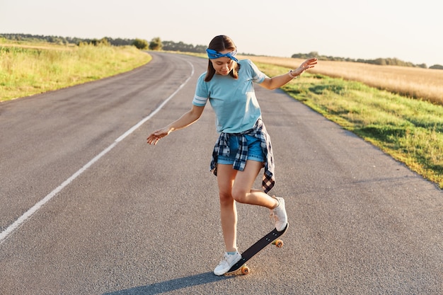 Outdoor-Aufnahme einer sportlichen Frau in Freizeitkleidung, die Spaß auf der Asphaltstraße mit Skateboard hat, verbringt gerne Zeit auf aktive Weise, gesunden Lebensstil, glücklichen Zeitvertreib.