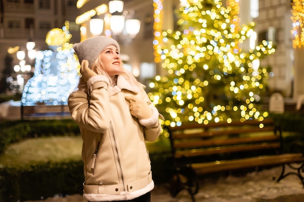 Outdoor-Foto des jungen schönen glücklichen lächelnden Mädchens, das Wunderkerzen hält und in der Straße aufwirft. Festlicher Weihnachtsmarkt im Hintergrund. Modell mit stilvollem Wintermantel, Strickmütze, Schal.