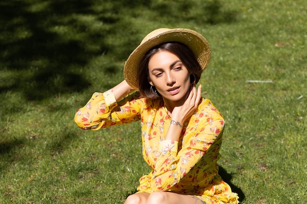 Outdoor-Modeporträt der Frau im gelben Sommerkleid und im Hut, die auf Gras im Park sitzen