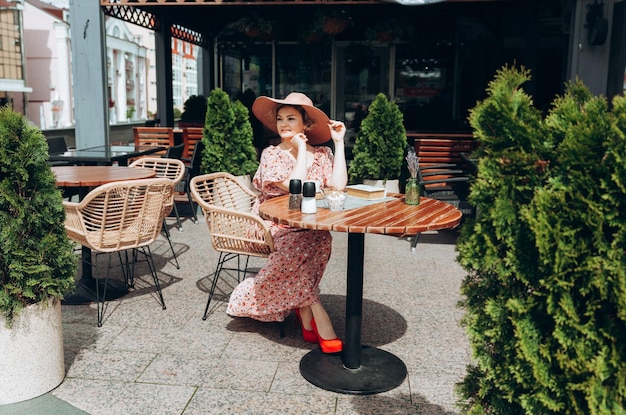 Outdoor-Modeporträt einer atemberaubenden Frau, die in einem Café sitzt. Ich trinke Kaffee und lese ein altes Buch, eine Frau in einem Kleid und einem Hut