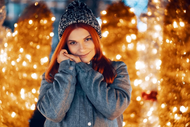 Outdoor-Nachtfoto eines jungen, schönen, glücklich lächelnden Mädchens, das festliche Dekoration genießt, auf dem Weihnachtsmarkt posiert, in der Straße der europäischen Stadt, mit gestrickter Beanie-Mütze