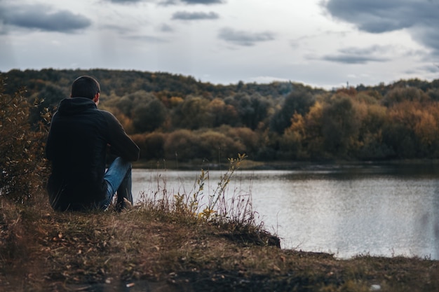 Outono nublado escuro. O cara sentado na margem do rio por trás. Solidão