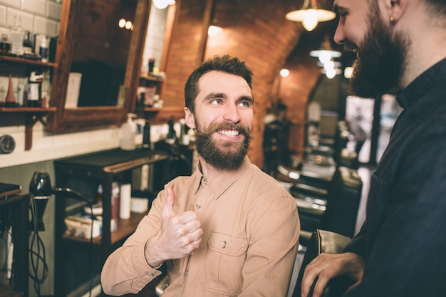 Outra foto de cliente feliz está olhando para o cabeleireiro e sorrindo. Ele está satisfeito com o resultado. Ele está mostrando o polegar grande até o cabeleireiro.