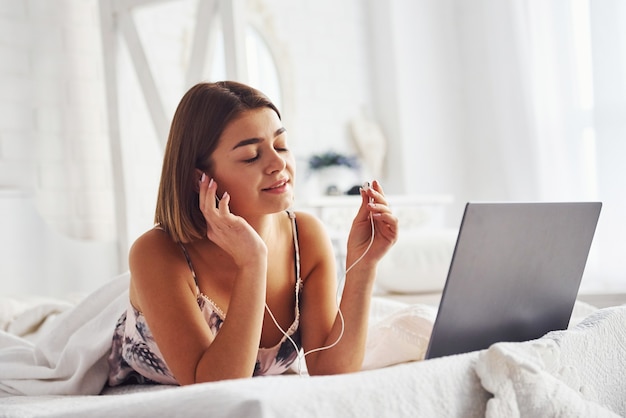 Ouvir música usando fones de ouvido. Linda garota deitada na cama com o laptop no quarto no seu tempo de fim de semana.