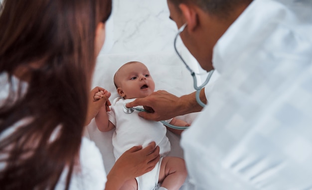 Ouvir usando o estetoscópio. Jovem pediatra está com o bebê na clínica durante o dia.
