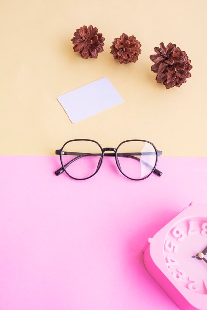 Ovale Brille auf dem Foto im minimalistischen Sommerstil auf pastellrosa und gelbem Hintergrund. Wecker , Kiefernblüten , Visitenkarten