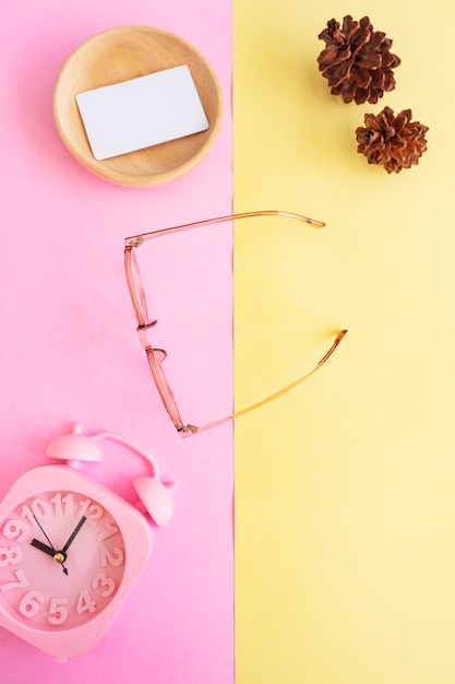 Ovale Brille auf dem Foto im minimalistischen Sommerstil auf pastellrosa und gelbem Hintergrund. Wecker , Kiefernblüten , Visitenkarten