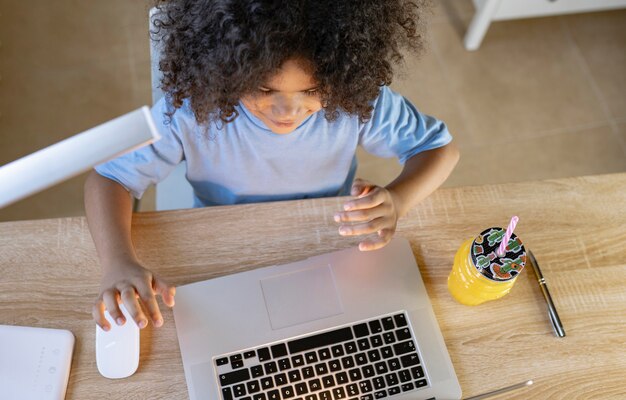 Overhead-Aufnahme eines Kindes, das zu Hause mit seinem Computer, einem Notebook und einem Orangensaft Hausaufgaben macht