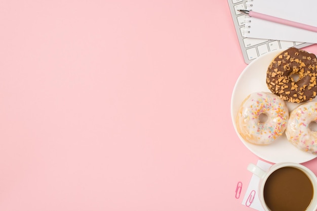 Overhead-Foto der Tastatur, Tasse Kaffee, Notizbuch mit Büroklammern und Teller mit glasierten Donuts, isoliert auf rosafarbenem Hintergrund mit leerem Raum