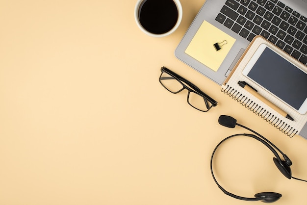 Overhead-Foto von grauem Laptop, Tasse Kaffee, Notizblock mit Stift, Brille, Telefon, Büroklammern, Kopfhörern und Aufklebern, isoliert auf dem beigen Hintergrund mit leerem Raum