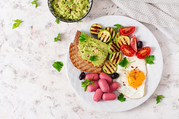 Ovo frito com mini linguiça tomate abobrinha grelhada azeitonas pretas e torradas com guacamole Café da manhã inglês Brunch Vista de cima