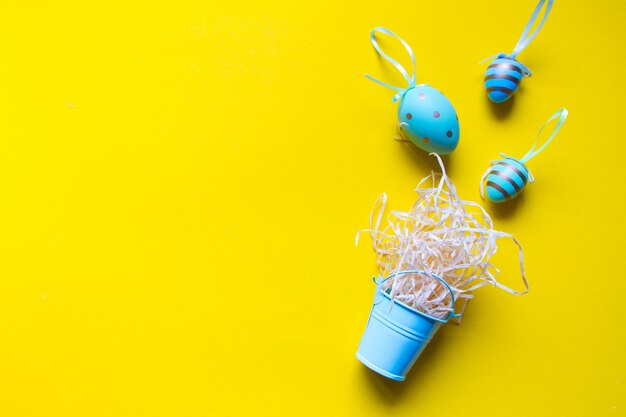 Ovos coloridos decorativos e um ninho de palha em um balde em miniatura sobre um fundo amarelo brilhante, fundo minimalista da Páscoa ou conceito