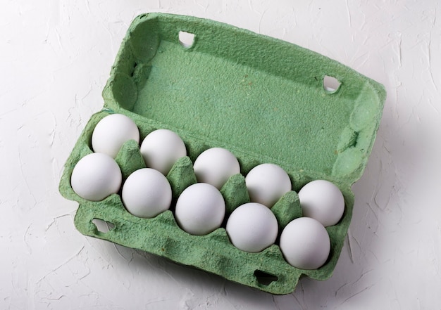 Ovos de galinha branca em uma caixa de papelão verde, em uma vista superior do plano de fundo texturizado branco.