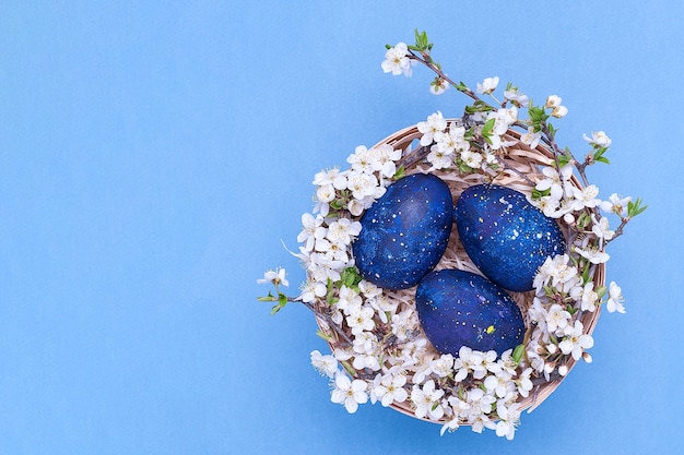 Ovos de Páscoa azuis em uma cesta com flores sobre fundo azul.