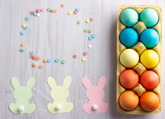 Ovos de páscoa coloridos, coelhinhos de papel e estrelas de açúcar na madeira branca