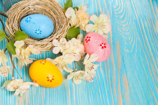 Ovos de páscoa coloridos em um ninho de palha e galho com flores sobre fundo azul de madeira