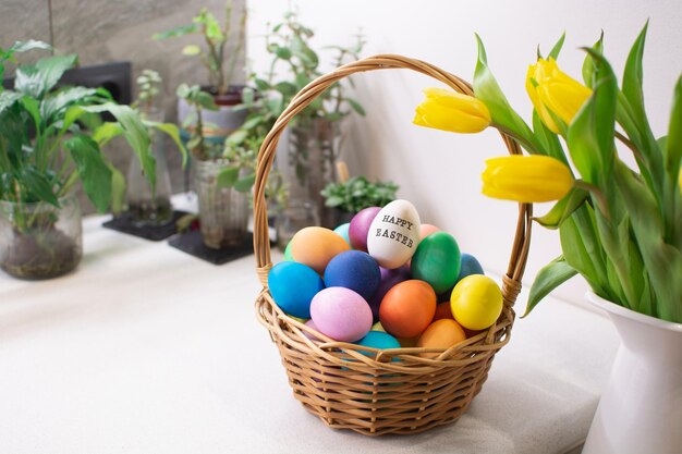 Ovos de páscoa coloridos na cesta de vime na mesa no interior da cozinha com tulipas