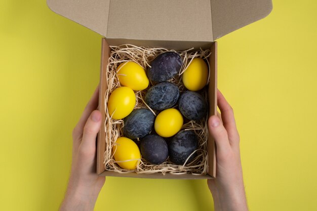 Ovos de Páscoa pintados nas cores azul e amarelo mármore em caixa de papelão na superfície amarela. Mãos segurando uma caixa.