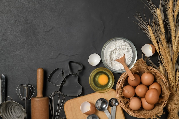 Ovos frescos e farinha de bolo com utensílios de cozinha para bolos na mesa preta,