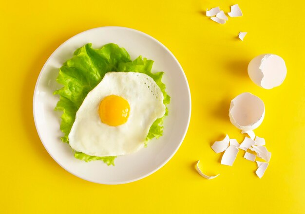 Ovos fritos com alface em uma casca de ovo de prato branco ao lado do prato em um fundo amarelo