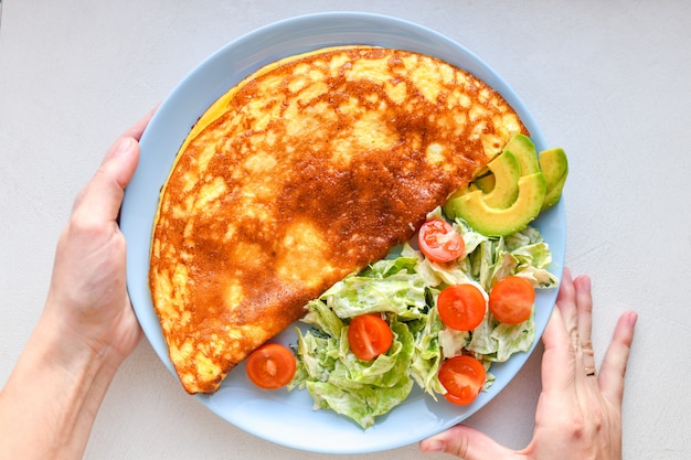 Ovos fritos com legumes. mãos no quadro. Omelete de legumes em um prato azul sobre uma mesa branca, vista superior