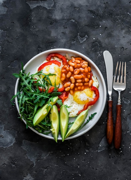 Ovos fritos com salada de rúcula doce pimenta vermelha abacate e feijão em molho de tomate um delicioso café da manhã em uma vista superior de fundo escuro