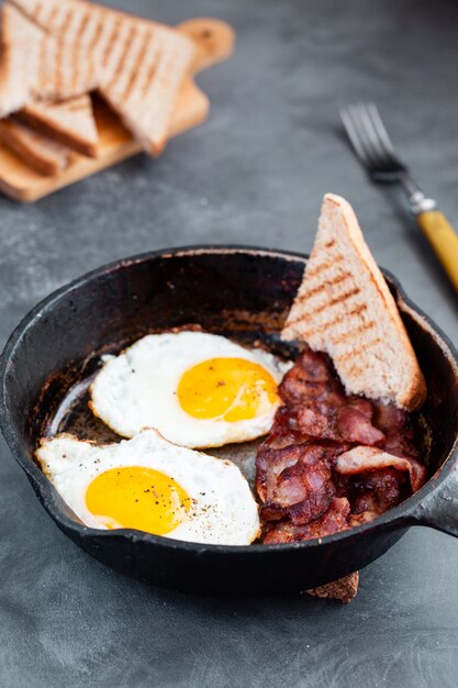 Ovos fritos em uma frigideira com tomate cereja seco, bacon e pão torrado no café da manhã em uma madeira rústica