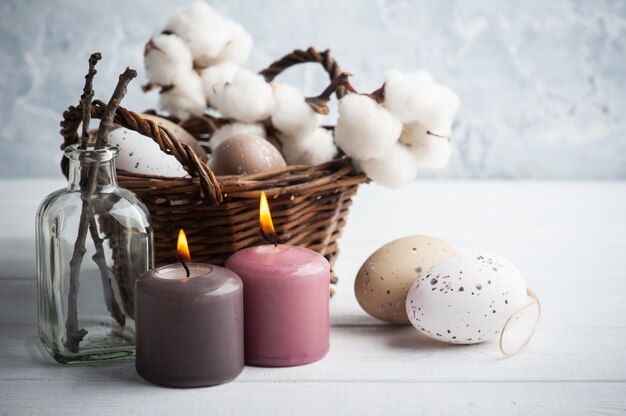 Ovos marrons, cesta de vime na composição rústica de Páscoa com velas acesas na mesa de madeira branca.