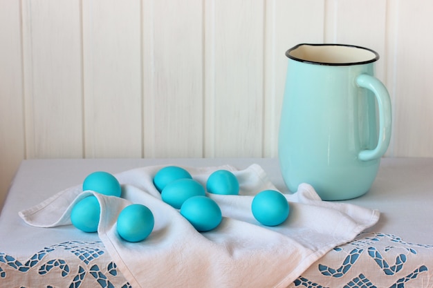 Ovos pintados de azul e um jarro esmaltado em uma mesa com uma toalha de mesa branca