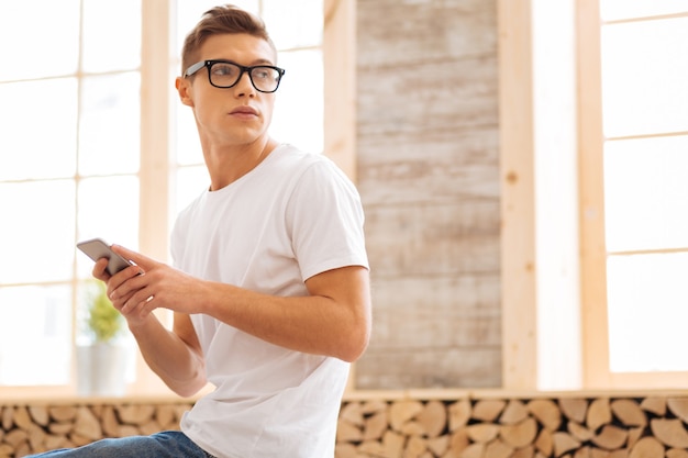 Foto oye. atractivo joven concentrado con gafas y sosteniendo su teléfono y mirando por encima del hombro