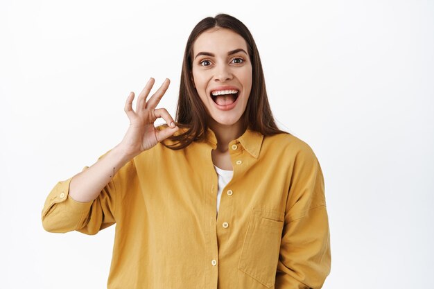 Oye, eso fue excelente. Mujer joven emocionada sonriendo asombrada, mostrando un buen gesto para alabar algo genial, viendo una actuación impresionante y hacer un cumplido, de pie sobre un fondo blanco.