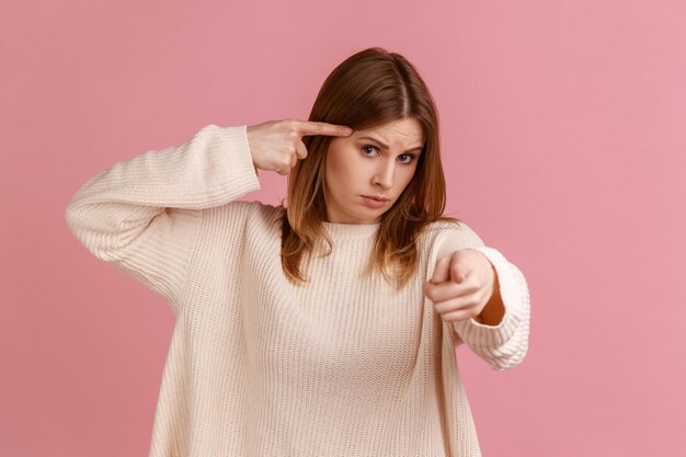 Oye, estás bromeando. Retrato de mujer rubia sosteniendo el dedo cerca de la cabeza y señalando con el dedo a la cámara, gesto estúpido, usando suéter blanco. Disparo de estudio interior aislado sobre fondo rosa.