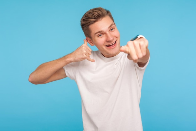 Oye, llámame Retrato de un joven apuesto y positivo con camiseta blanca haciendo gestos telefónicos y señalando a la cámara esperando comunicación en un estudio interior aislado de fondo azul