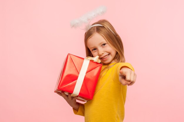 Oye, este regalo para ti. Retrato de feliz niña de jengibre con halo angelical abrazando la caja envuelta y apuntando a la cámara, eligiendo al afortunado ganador para dar un bono, regalo. tiro del estudio de interior, aislado