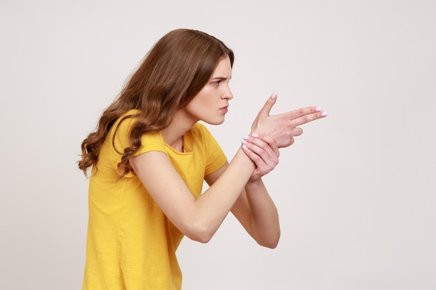 ¡Oye, te mataré! Vista lateral de una joven con una camiseta informal amarilla que apunta con pistolas de dedo, fingiendo disparar, amenazando con un gesto de pistola de mano. Disparo de estudio interior aislado sobre fondo gris.