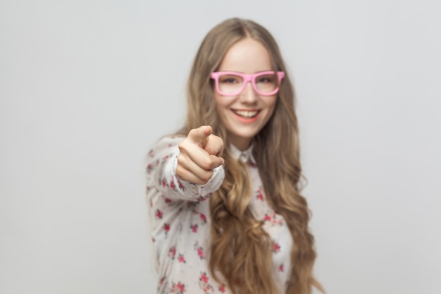 ¡Oye tú! ¡Genial! Chica adolescente, señalando con el dedo a la cámara y sonriendo con dientes. Foto de estudio, aislado sobre fondo gris